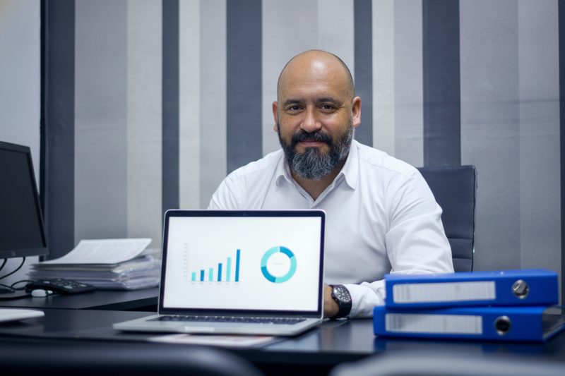 El mismo empresario, tranquilo, con el escritorio ordenado y un panel interno de ejemplo en su laptop.